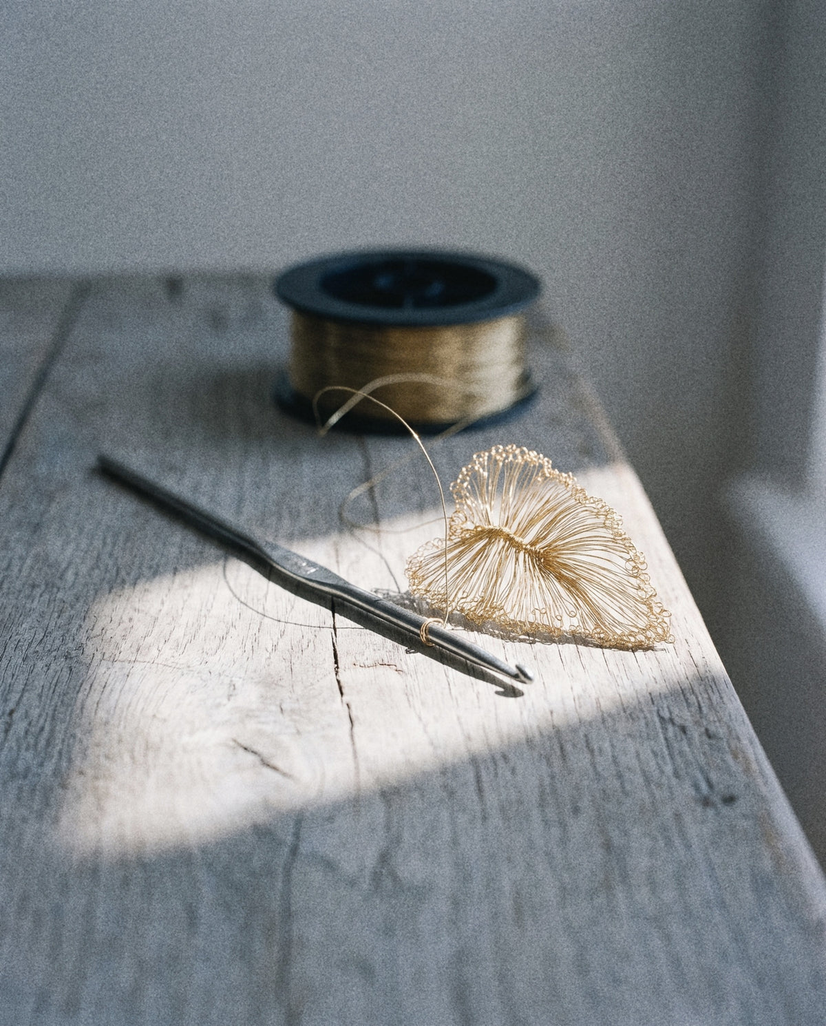 A steel crochet hook and an unfinished leaf of gold-tone wire on a wooden table, with the spool behind