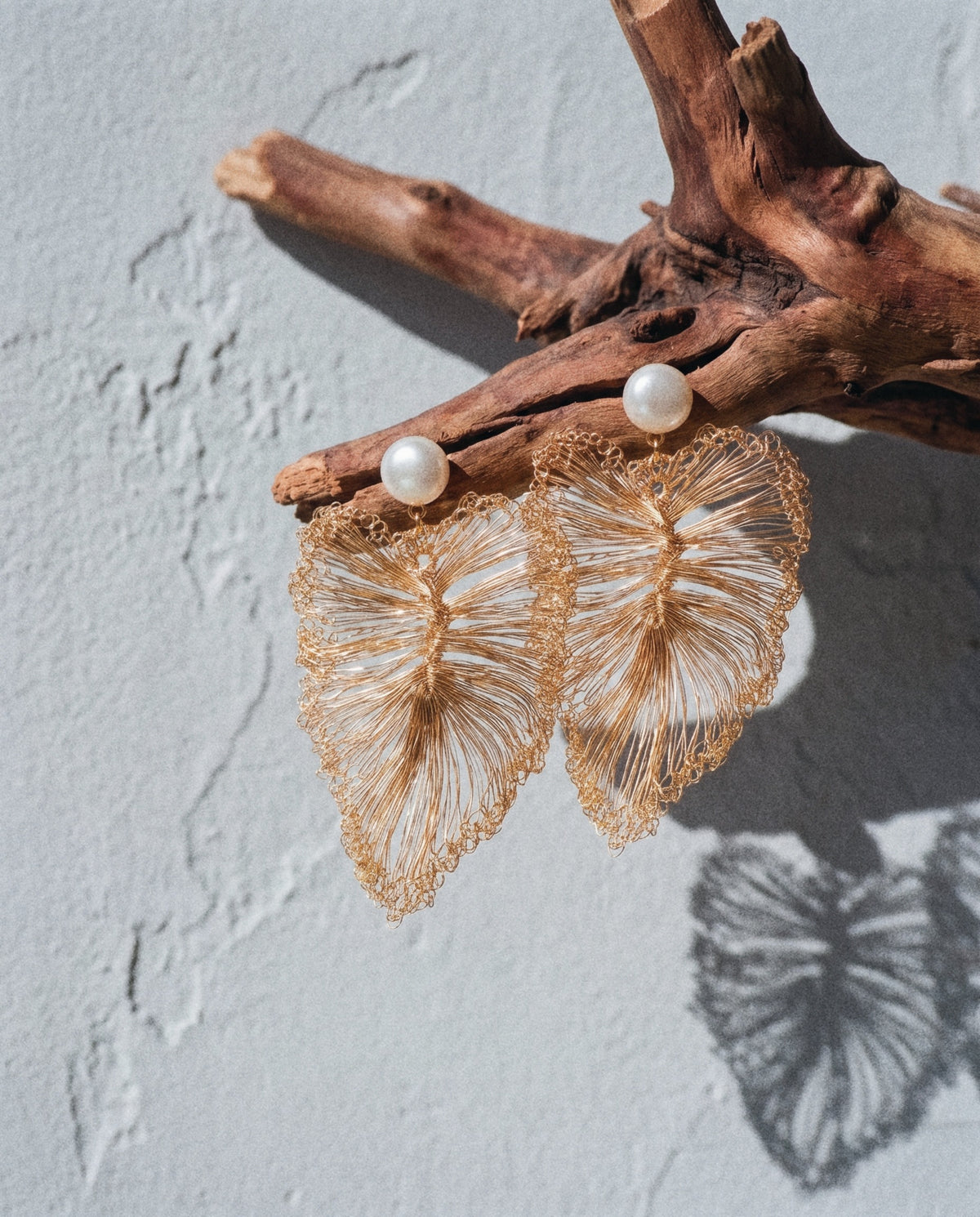 A pair of crocheted wire leaf earrings with pearl studs hanging from a piece of driftwood